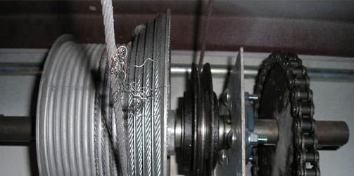 Close-up of a garage door cable drum with frayed cable and chain drive.