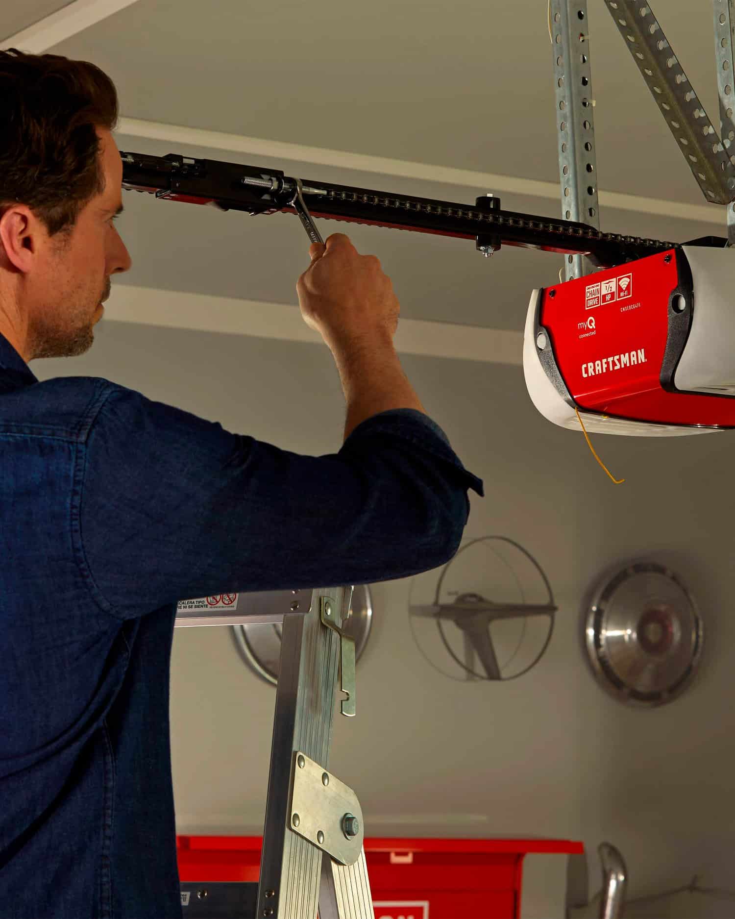 Man on a ladder adjusting a garage door opener with a wrench.