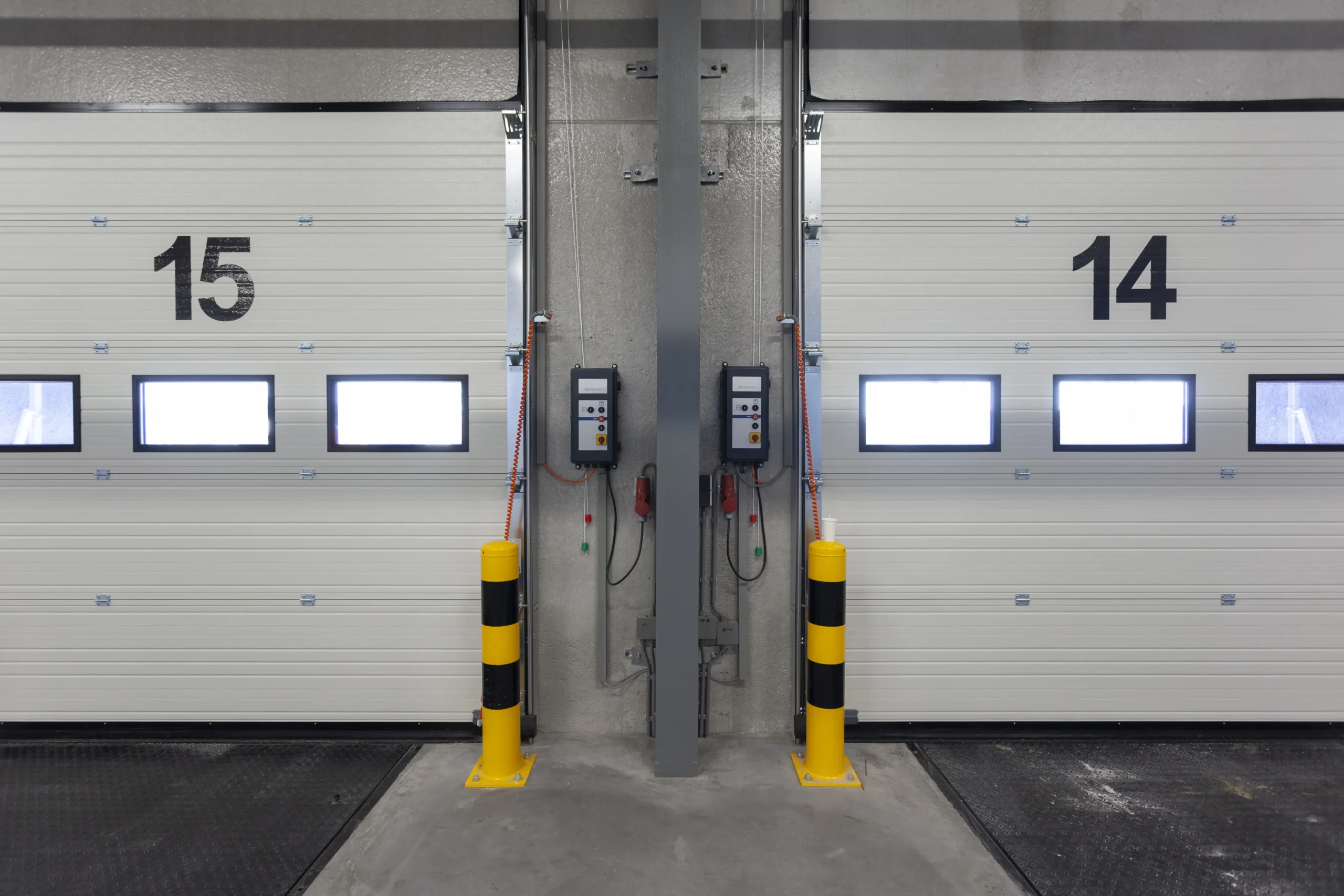 Two white loading dock doors, numbered 15 and 14, with yellow and black bollards.