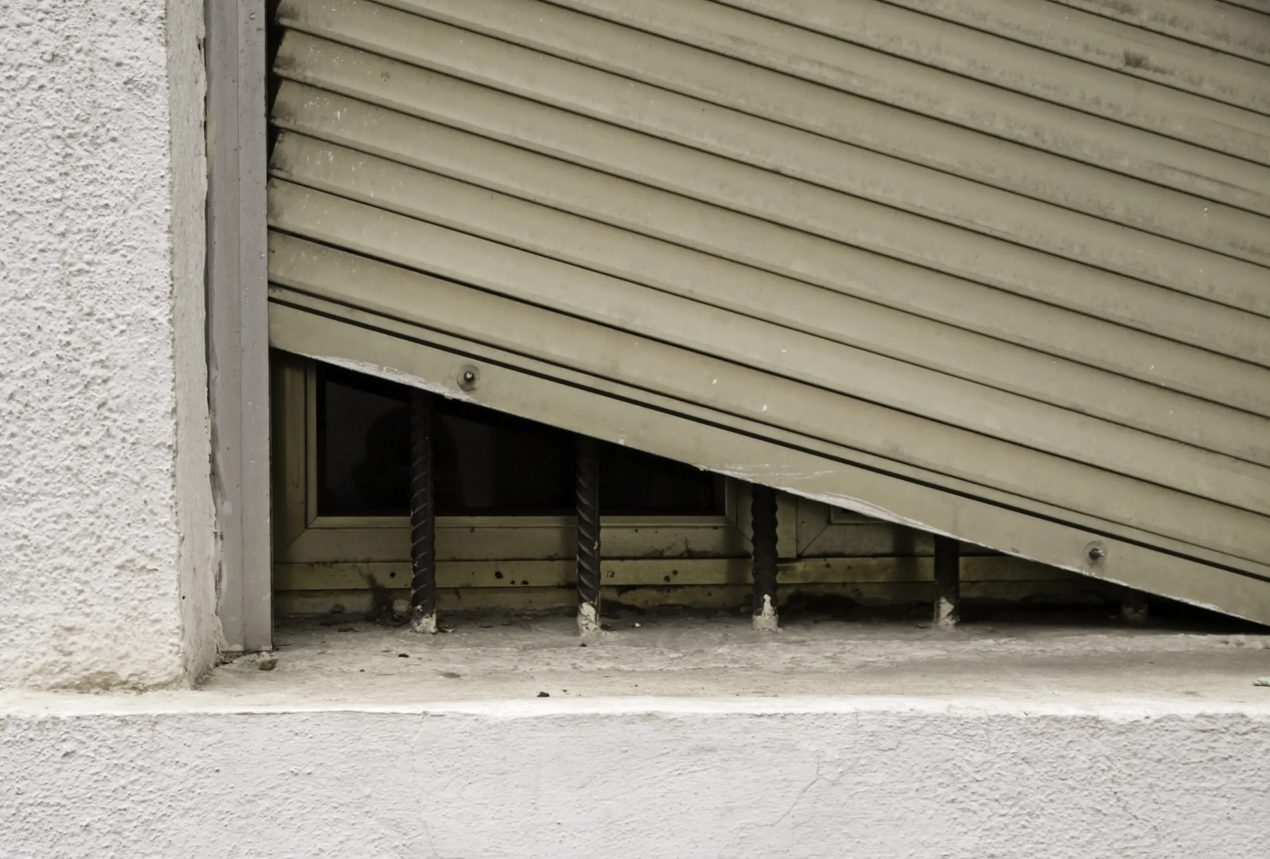 Broken shutter on a window with security bars and a dark interior.