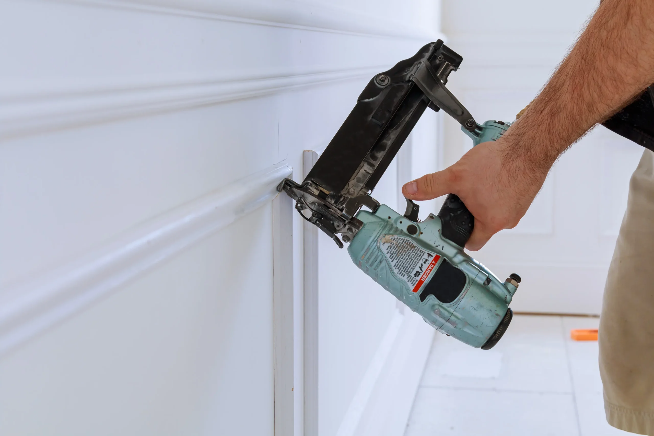 Close-up of a person using a nail gun on white wall trim.