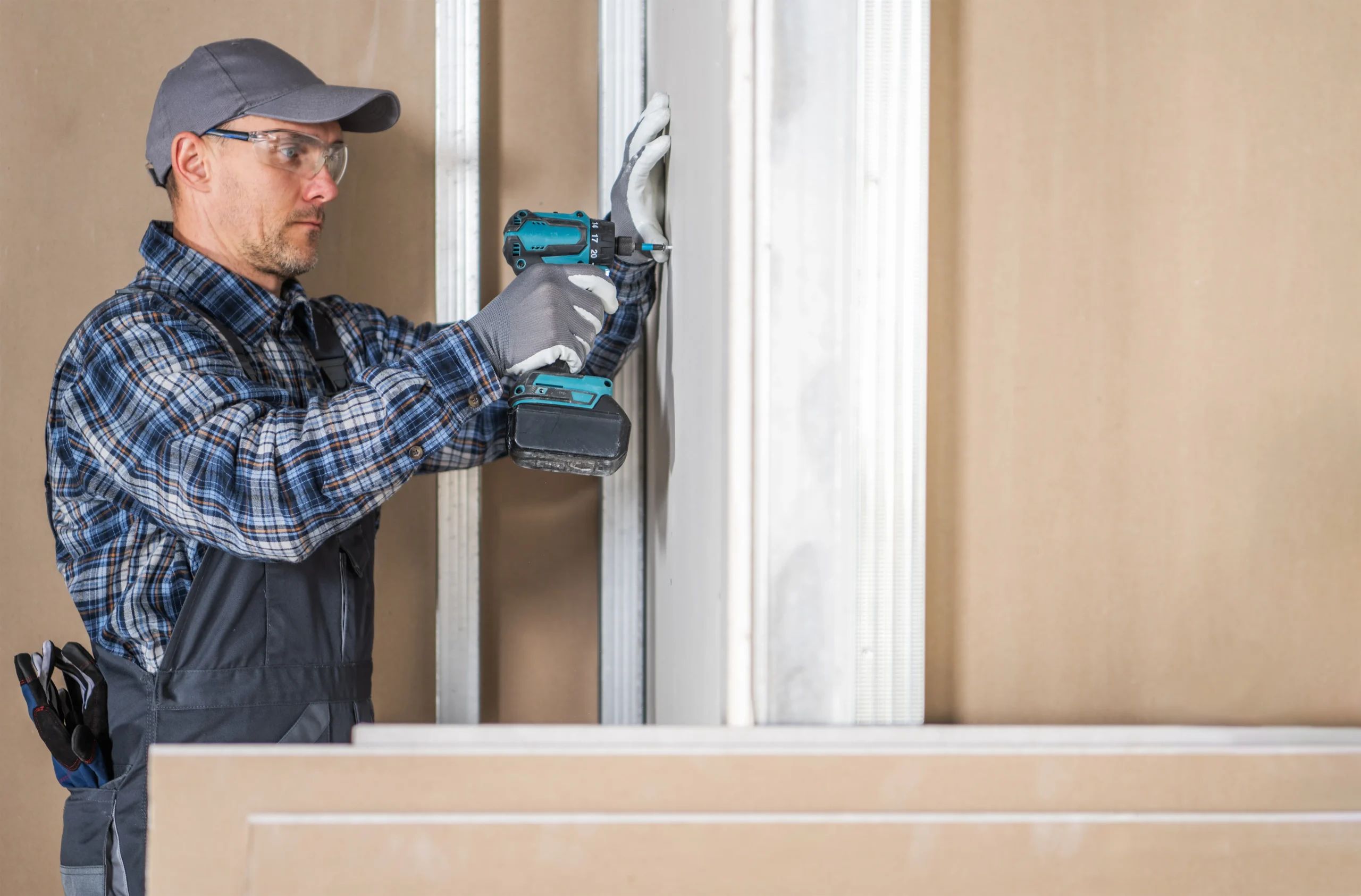 Man in safety glasses and cap using a drill on a wall.