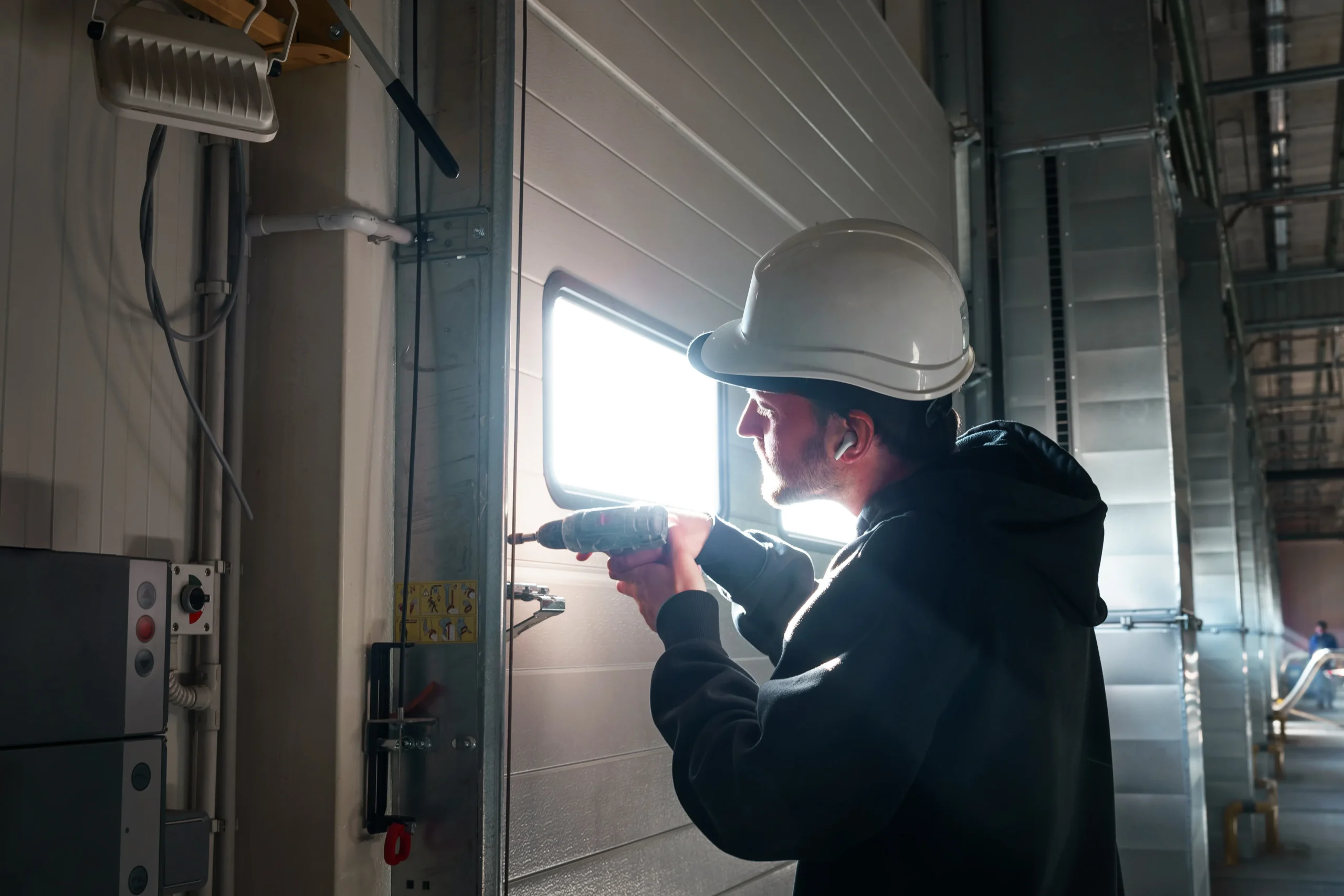 Man in hard hat drilling into a garage door panel with a window.
