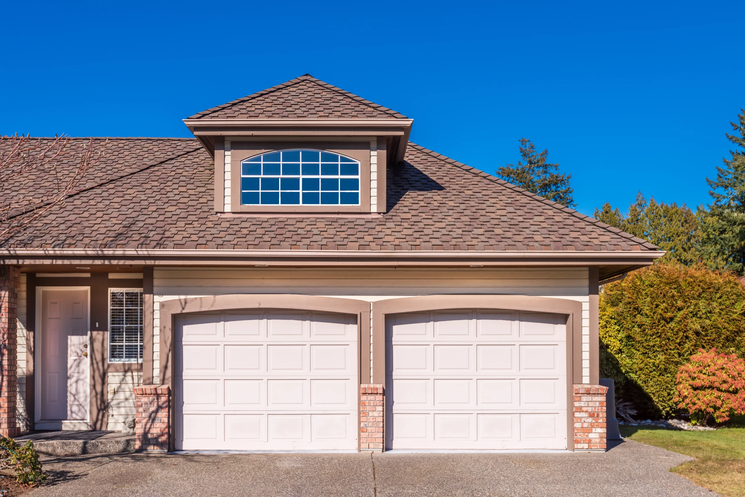 Two white garage doors on a house with a brown roof and blue sky.