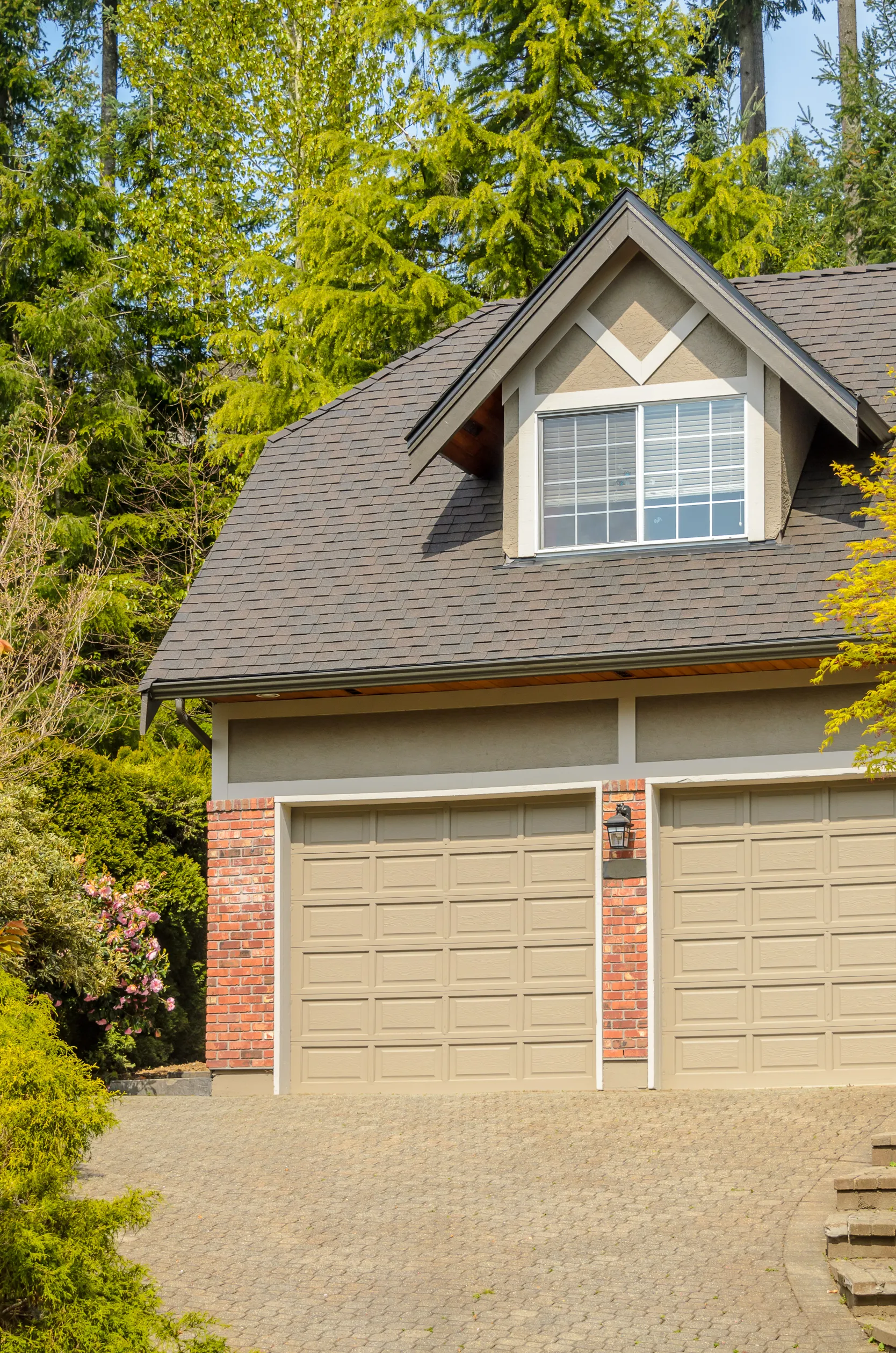 Two beige garage doors on a house with a dormer window and green trees.