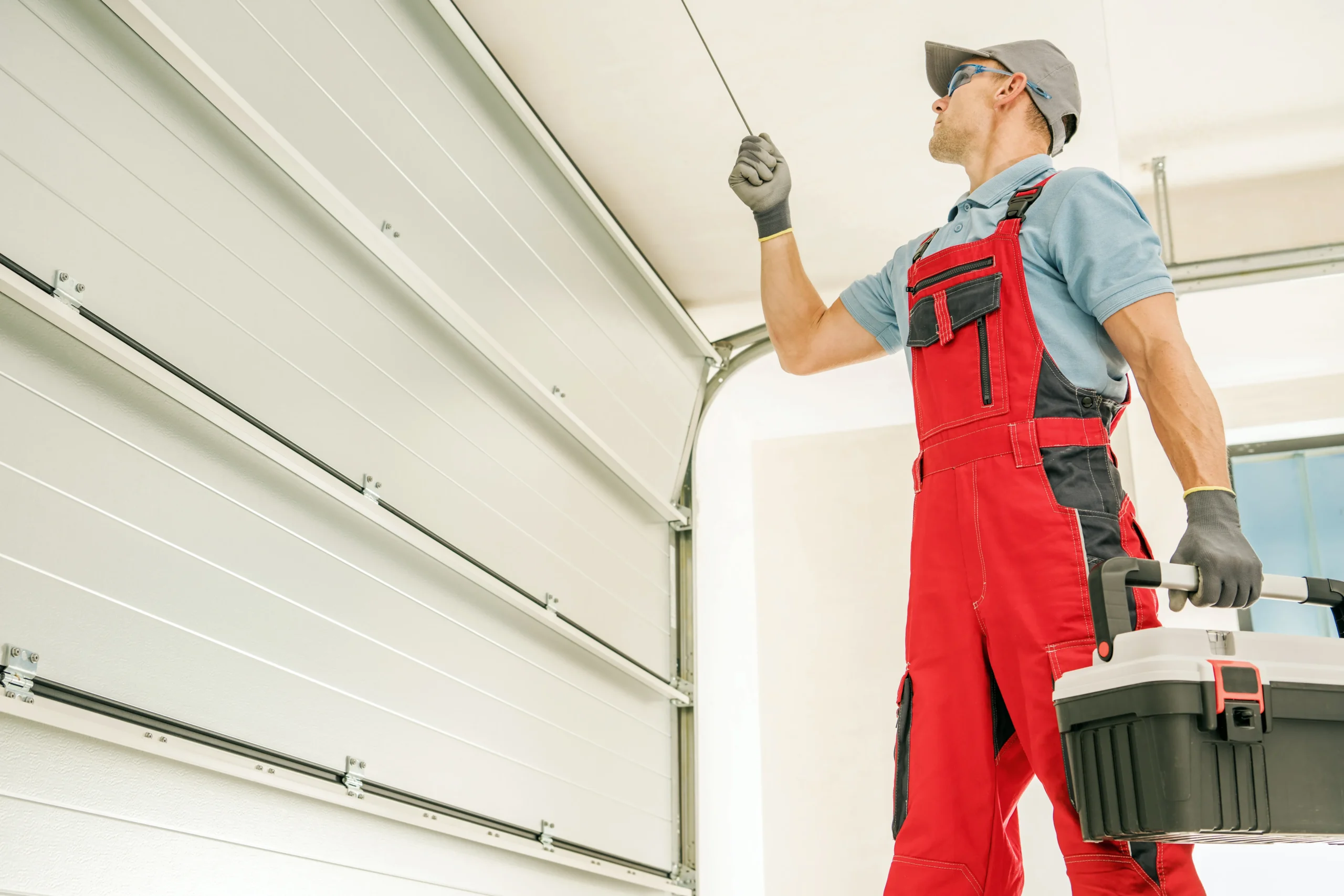Man in red overalls and grey cap inspecting a garage door mechanism.
