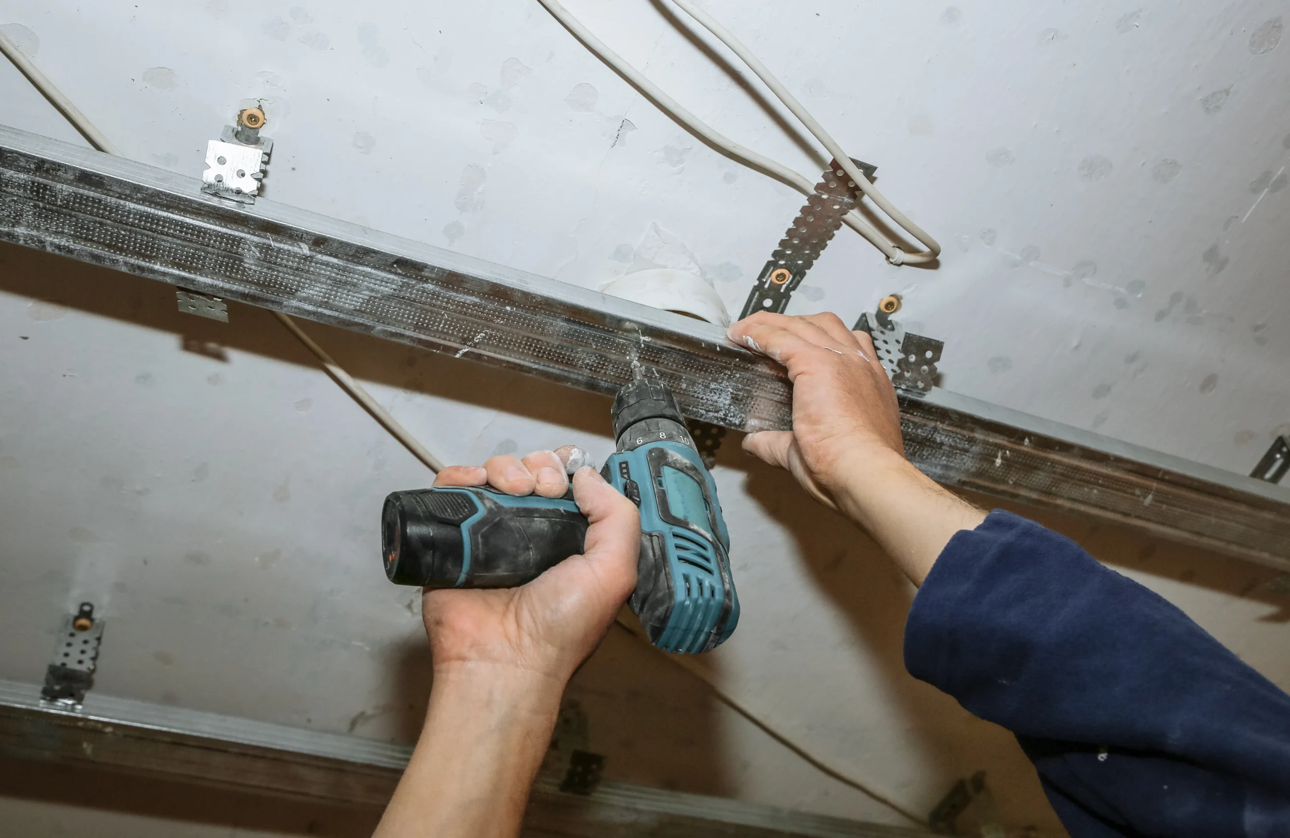 Hands using a drill to install metal framing on a ceiling.