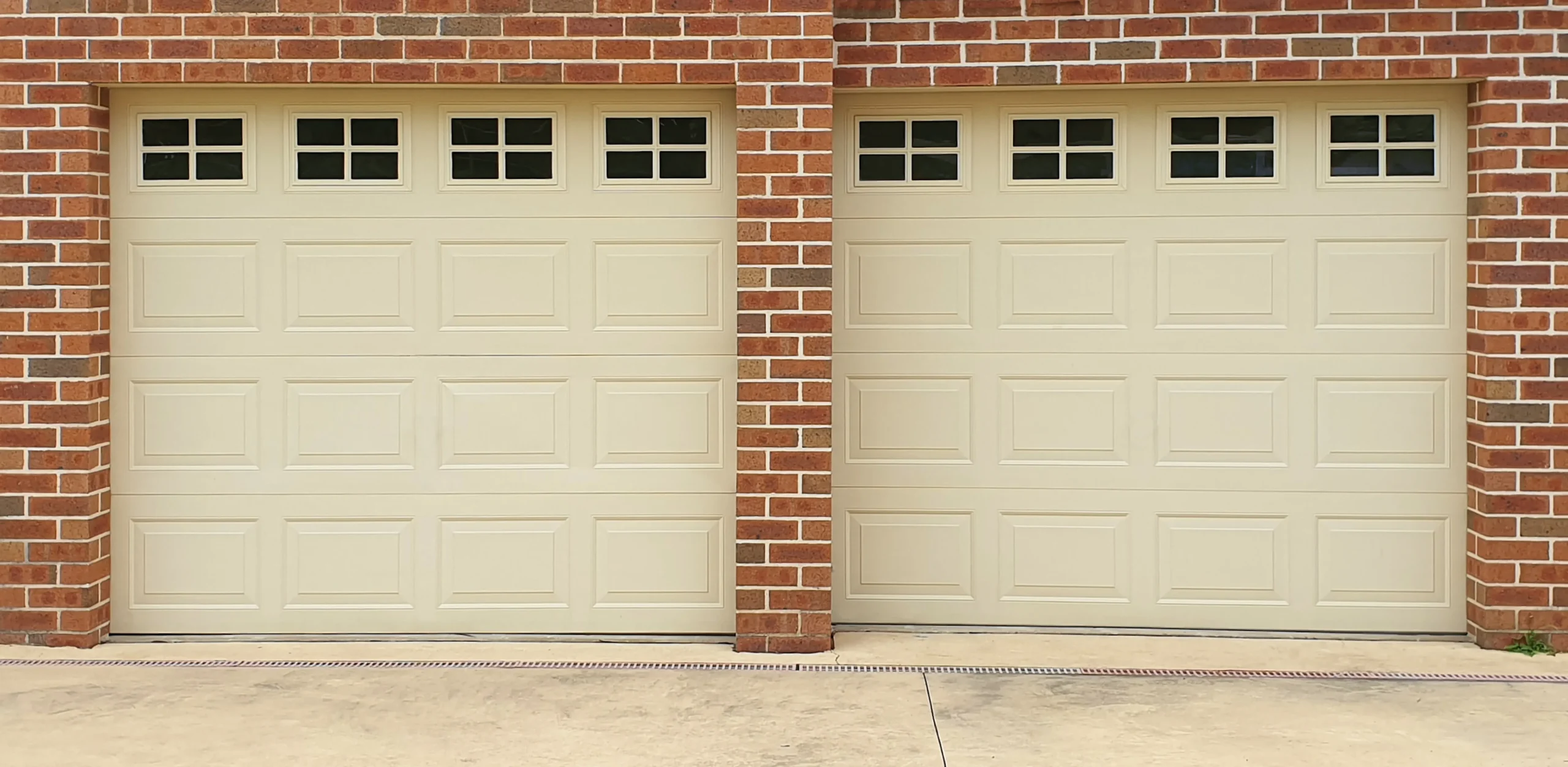 Two beige garage doors with windows on a brick house.