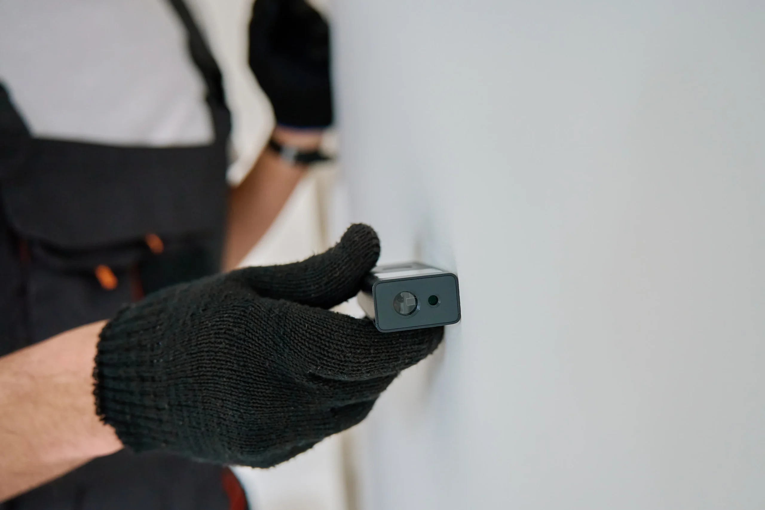 Person in black gloves holding a small black device against a white wall.