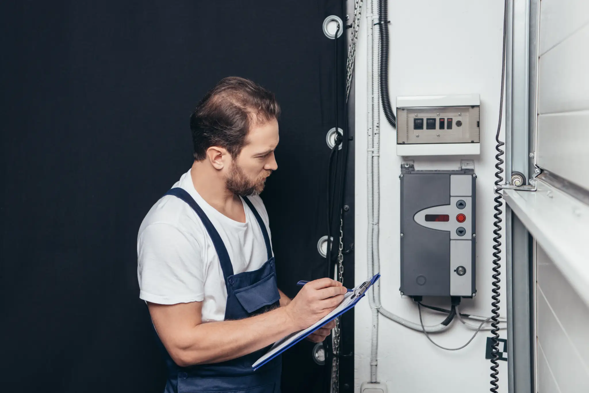 Man in overalls writing on a clipboard next to a garage door control panel.