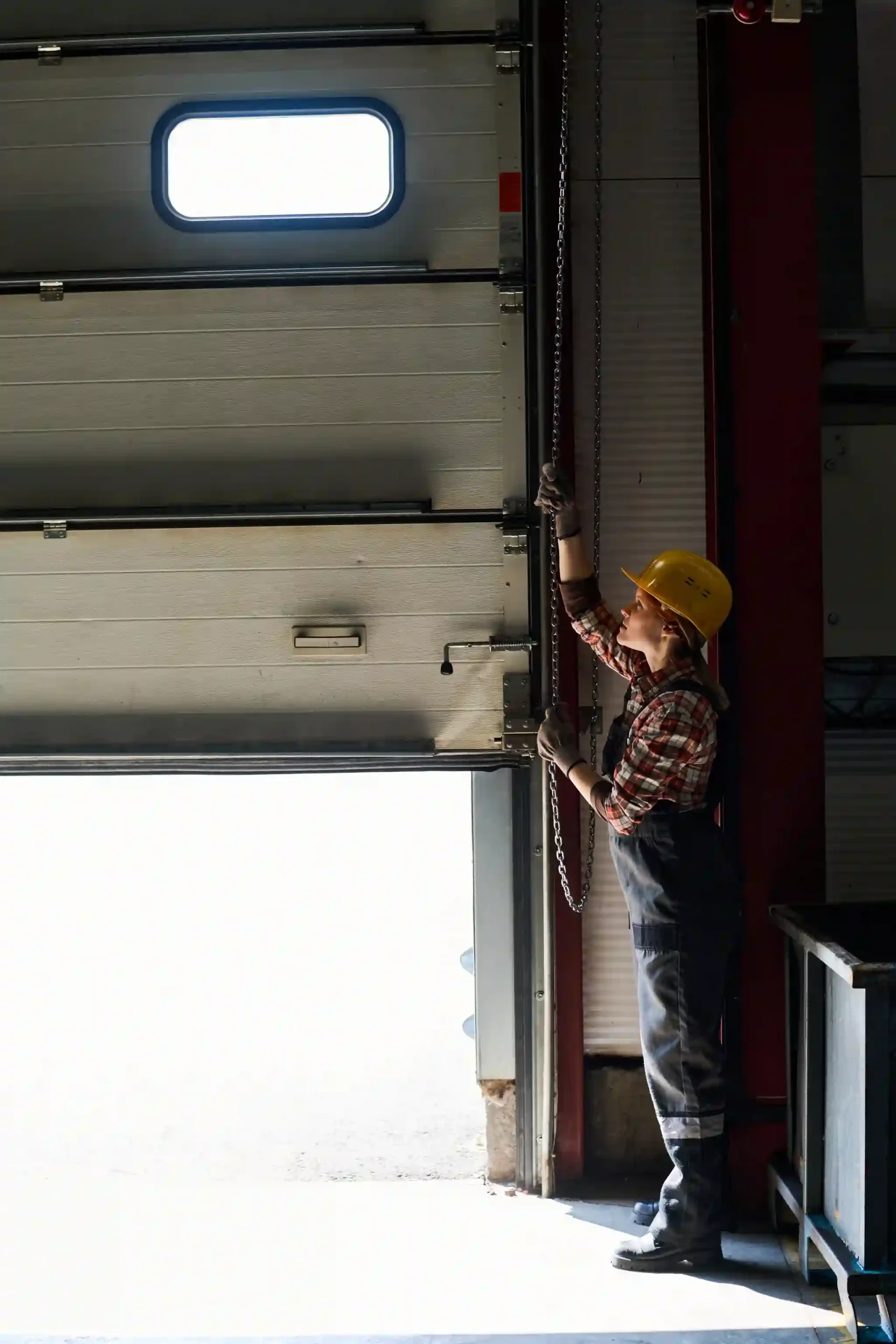 Woman in hard hat and overalls opening a large garage door with a chain.
