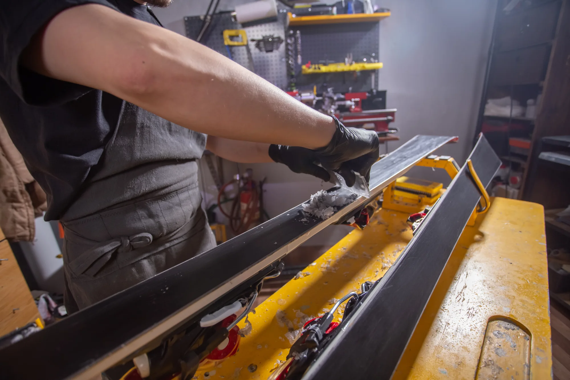 Person wearing black gloves scraping wax off skis on a yellow workbench.
