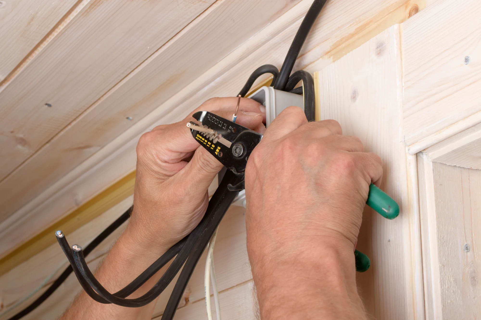 Hands stripping wires with a tool during electrical installation on a wooden wall