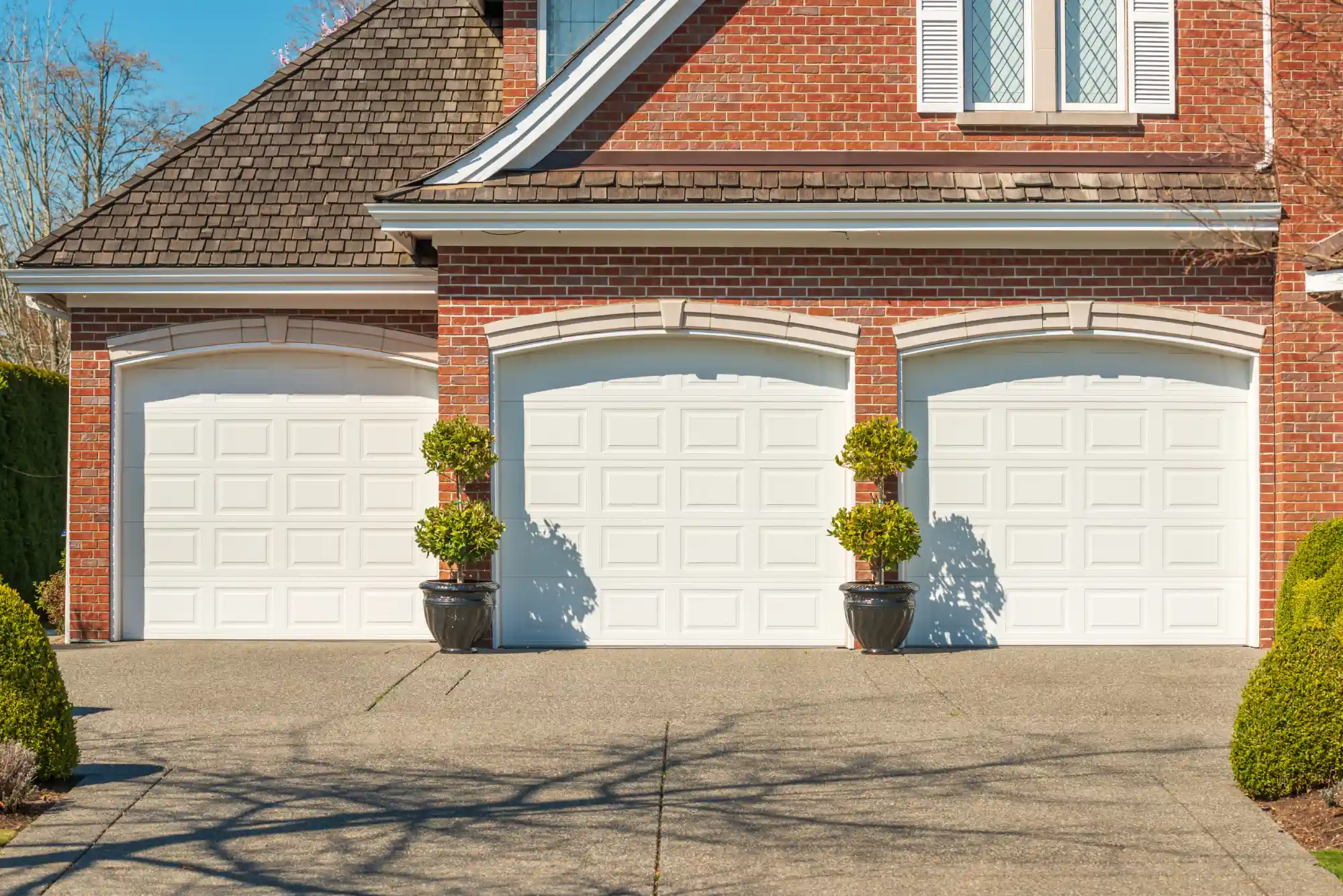 Three white garage doors on a brick house with a paved driveway.