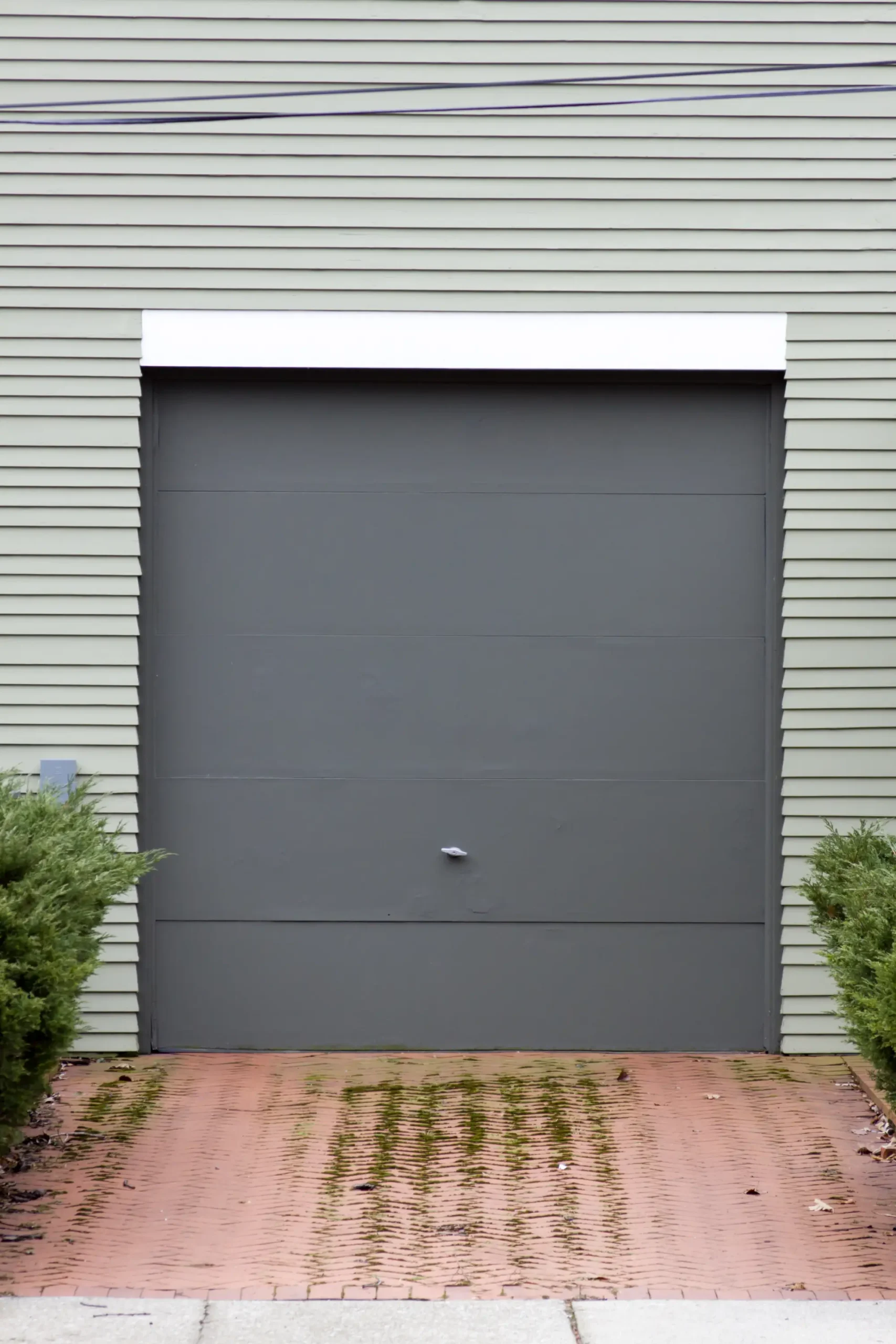 A gray garage door with a white trim, green siding, and brick driveway.