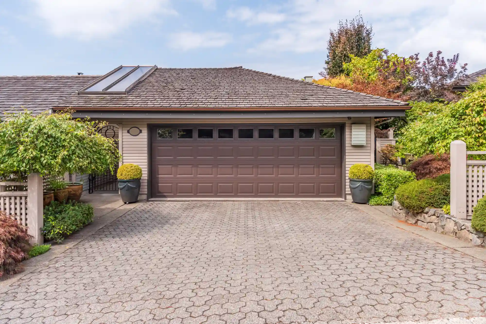 Brown garage door with a patterned driveway and lush landscaping.