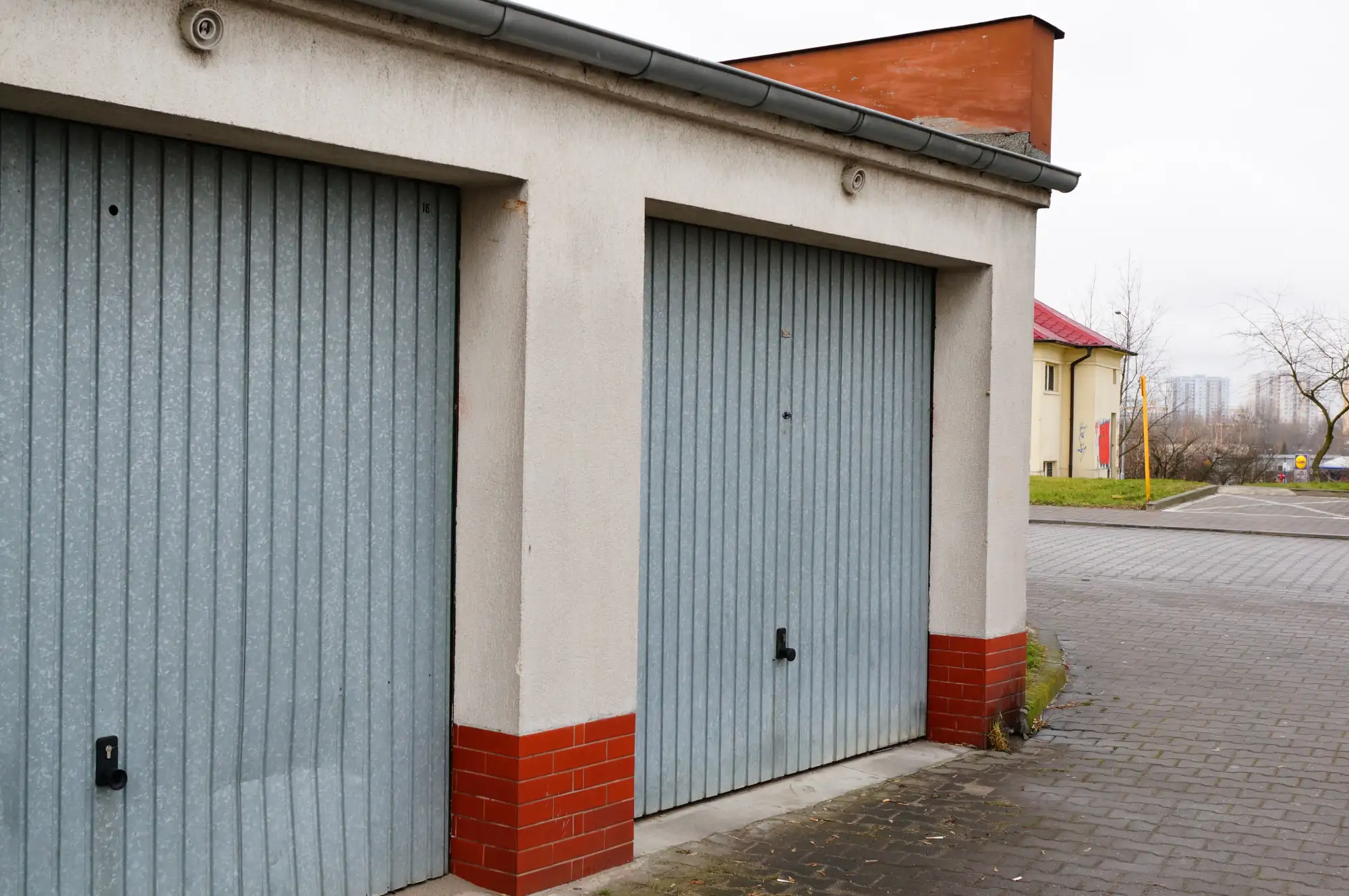 Two light blue garage doors with a paved driveway and distant buildings.