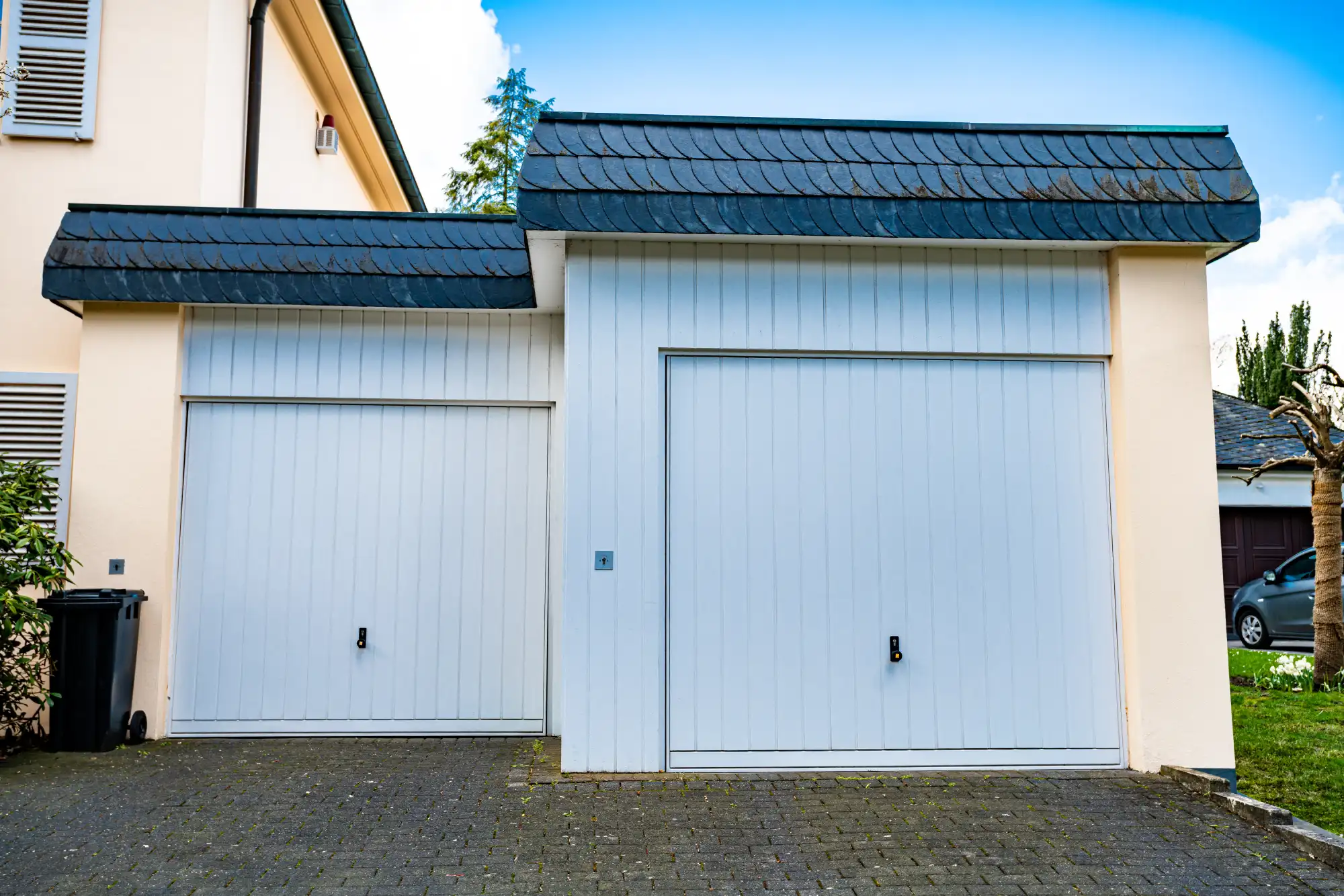 Two white garage doors on a light-colored house with a dark roof.