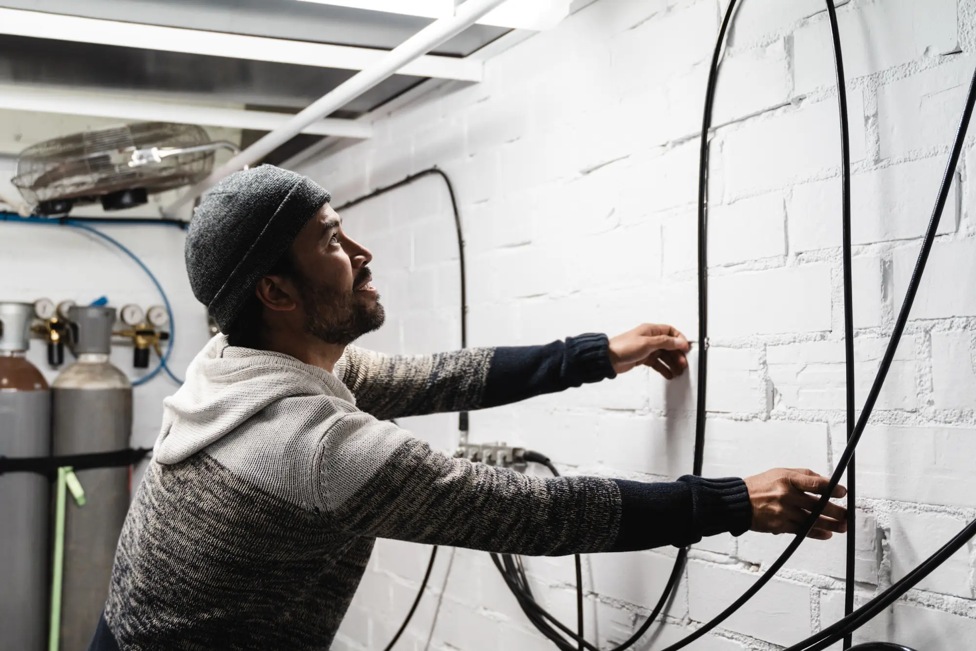 Man in a beanie and sweater adjusting wires on a white brick wall