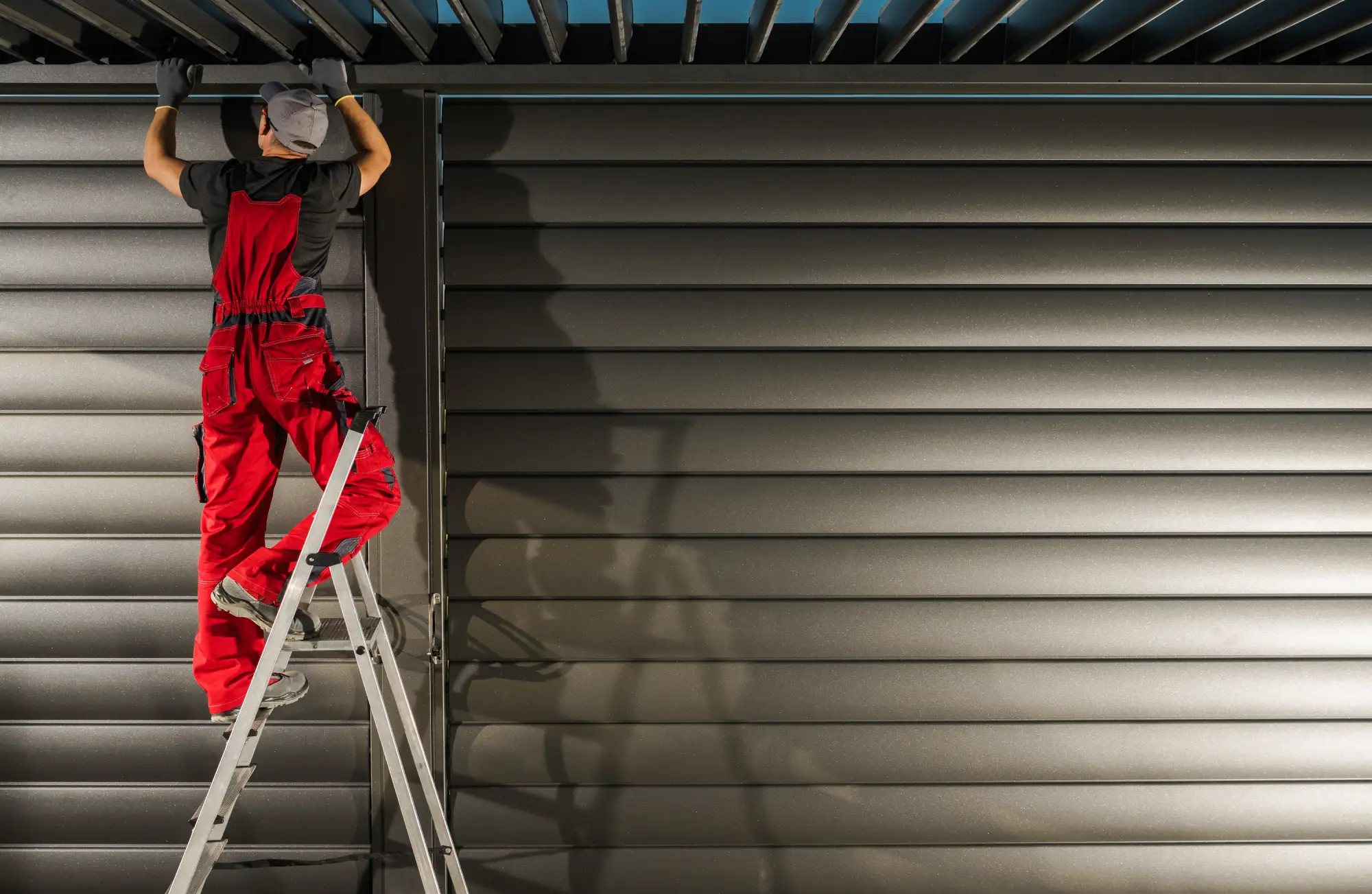 Man in red overalls on a ladder installing a dark gray louvered roof.